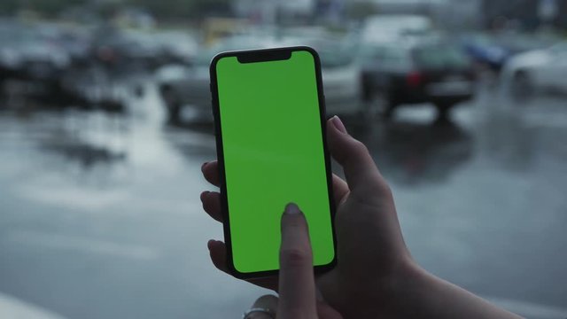 Los Angeles, California - 5 April 2019: Woman hand holding use touch vertically smart phone cellphone with green screen background blurred rain parking cars app cellphone outside rain trees raindrops
