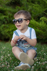 Cute little boy sitting on grass field wearing sunglasses. Adorable caucasian child   playing outdoor in summer park.