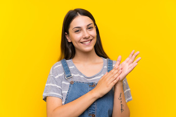 Young woman in dungarees over isolated yellow background applauding