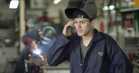 4K Young mechanic wearing a welding helmet talking on phone in garage workshop