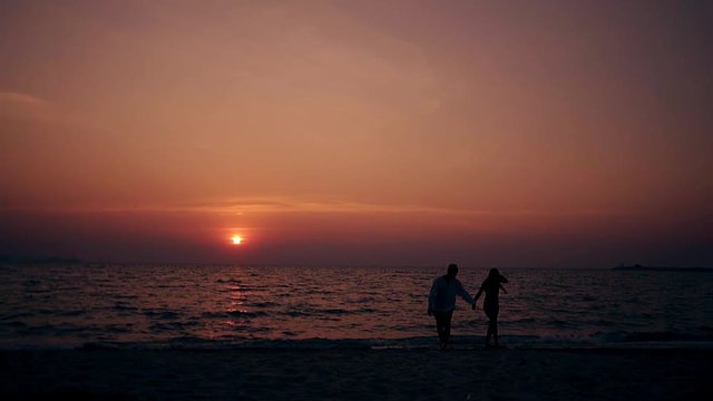 silhouette of young couple joining hands and walking along empty ocean beach against red sun slow motion