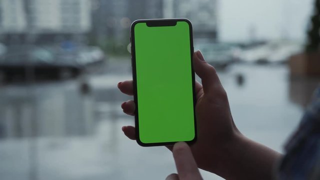 Los Angeles, California - 5 April 2019: Close up view woman hands use holding vertically smart phone cellphone with green screen background rain parking cars weather app outside rain trees raindrops