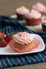 Homemade strawberry cupcakes in red and blue paper cups on cooling rack.  One isolated in front on blue paper towel.  Fresh strawberries scattered around.