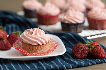 Homemade strawberry cupcakes in red and blue paper cups on cooling rack.  One isolated in front on blue paper towel.  Fresh strawberries scattered around.