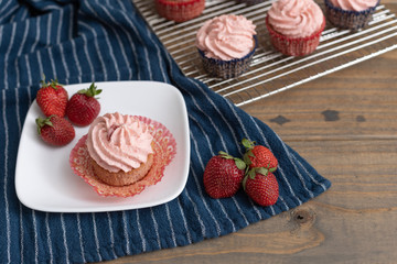 Homemade strawberry cupcakes in red and blue paper cups on cooling rack.  One isolated in front on blue paper towel.  Fresh strawberries scattered around.