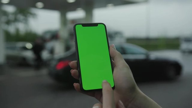 Los Angeles, California - 5 April 2019: Slow motion woman hand holding vertically smart phone cellphone with green screen background rain parking cars gas station outside rain trees raindrops