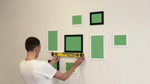 Young man corrects photo frames by building level. Photo frames hang on a wall.