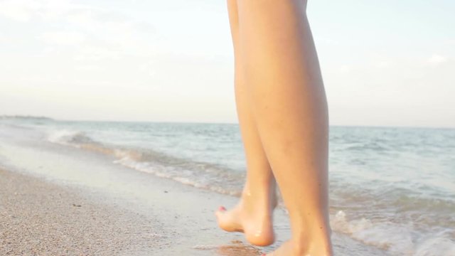 Point of view of young woman stepping at the golden sand at sea beach. Female legs walking near ocean. Bare foot of girl going on sandy shore with waves. Summer vacation or holiday Slow motion Closeup