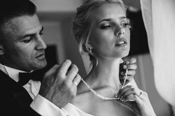 A groom is putting a necklace on a bride's neck. Black and white image.