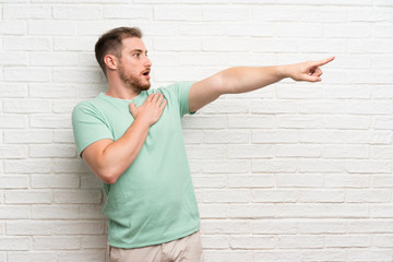 Blonde man over brick wall pointing finger to the side