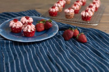 Homemade mini strawberry cupcakes in red and white paper liners.  Some isolated on blue plate with fresh strawberries.  Some cooling on rack in background.  Blue kitchen towel on wood table.