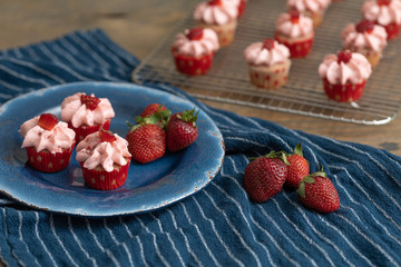 Homemade mini strawberry cupcakes in red and white paper liners.  Some isolated on blue plate with fresh strawberries.  Some cooling on rack in background.  Blue kitchen towel on wood table.
