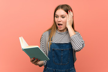 Young woman with overalls over pink wall surprised while enjoying reading a book