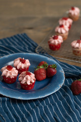 Homemade mini strawberry cupcakes in red and white paper liners.  Some isolated on blue plate with fresh strawberries.  Some cooling on rack in background.  Blue kitchen towel on wood table.