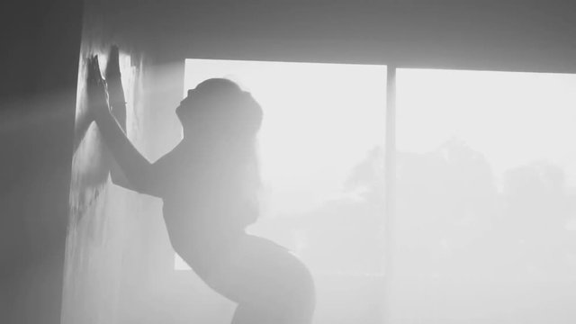 Silhouette of beautiful sensual woman in body suit dancing in hazy studio over window view background - black and white video
