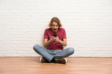 Blonde man sitting on the floor surprised and sending a message