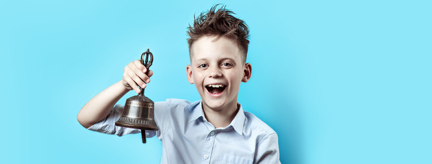 a happy boy in light shirt goes to school. He has a bell in his hand, which he rings and smiles.