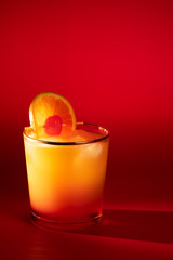 Tequila sunrise in a glass with golden rim garnished with orange slice and cherry. Red background, high resolution