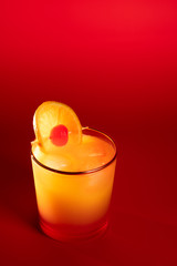 Tequila sunrise in a glass with golden rim garnished with orange slice and cherry. Red background, high resolution