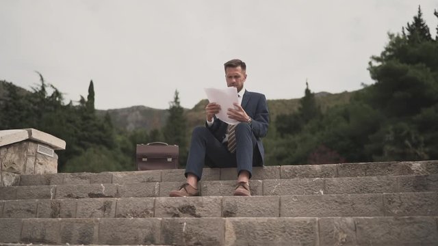 Businessman sits on the top step and scrolls through contracts, reads documents. Slow motion