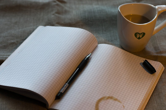 refiner from a cup of coffee on the notebook with open gel pen and cup of milk cofee on the flax backgruond on the table ander the lump light