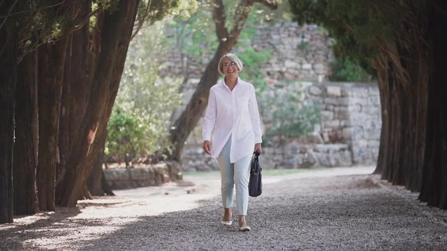 Confident senior business woman walking in city park. Slow motion