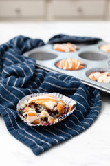 Freshly-baked lemon-glazed blueberry muffins in a muffin tin.  One isolated in front broken open.  Blue Kitchen towel on white countertop.