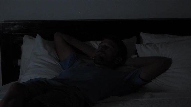 man dressed t-shirt shorts watching tv at night in a hotel lying on a bed.
