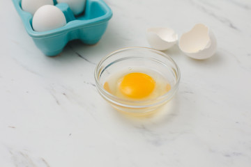 White eggs in turquoise egg tray.  One egg cracked open into clear bowl on white marble countertop.