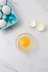 White eggs in turquoise egg tray.  One egg cracked open into clear bowl on white marble countertop.