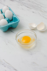 White eggs in turquoise egg tray.  One egg cracked open into clear bowl on white marble countertop.