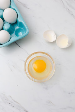 White eggs in turquoise egg tray.  One egg cracked open into clear bowl on white marble countertop.
