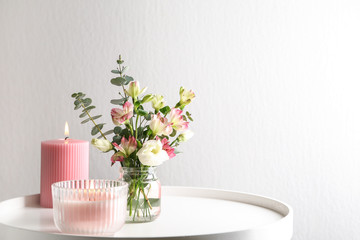 Stylish tender composition with burning candles and flowers on white table against light...