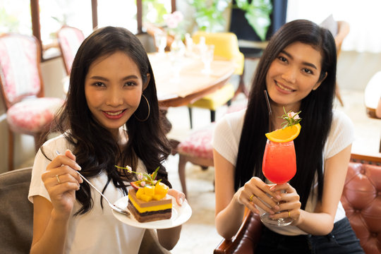 Two Asian Sisters take photo and taste delicious sweet Cake and Fruit Juice, Best friends smile together for beautiful design cake and smoothies refreshment in boutique restaurant cafe during travel
