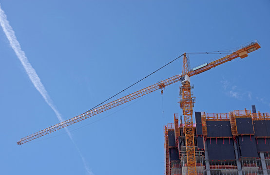 View of a yellow skyscraper construction crane under blue sky with a jet trail  - Powered by Adobe