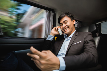 busy asian businessman calling by phone and using tablet on his way to meeting while sitting on passenger seat of his car