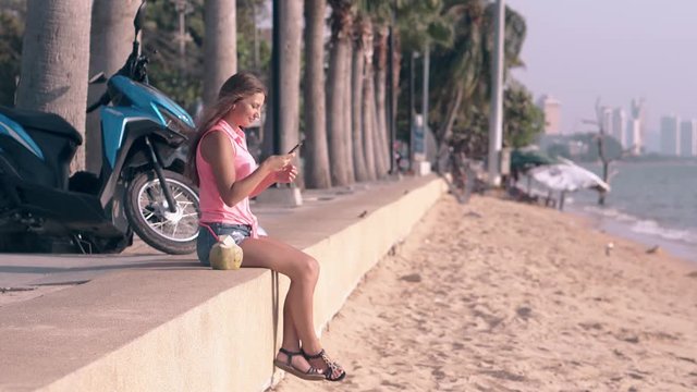young girl with long dark hair in bright pink top and denim shorts sits on concrete parapet and makes selfie on beach