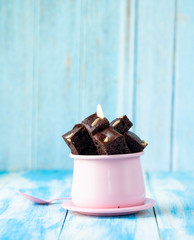Piece of brownie almond in pink cup with pink spoon and dry flower on blue wood table and blue background.