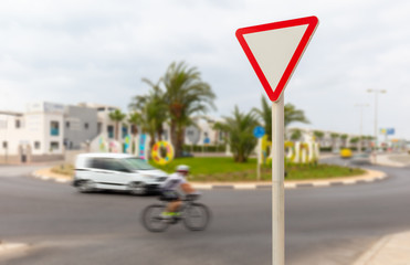A car and a road cyclist are in the grassy and palm-planted roundabout. A triangular red and white sign means pay attention.