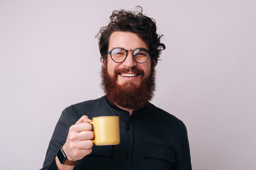 Photo of some cheerful bearded guy wearing glasses, loooking at camera and holding a mug with coffee, over isolated background
