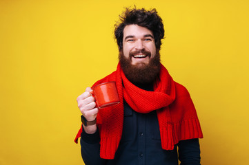 Photo of handsome bearded man,  wearing red scarf looking at camera with toothy smile and holding a mug with hot drink