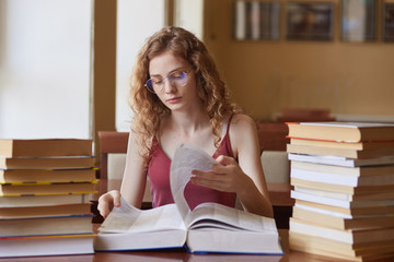 Image of thoughtful hard working young girl turning over pages of huge book, searching for proper information for study project, doing home assignment carefully. Student and deadline concept.