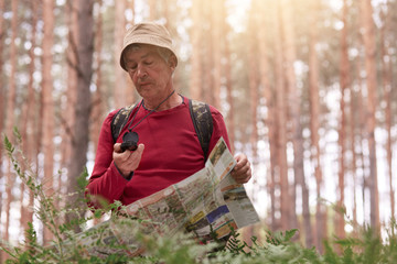 Image of eldery man hiking and looking at compass and map for direction for trip in forest, senior male wearing casual outfit with backpack being lost in wood. Traveling and active recreation concept.