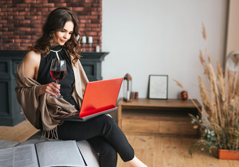 Young businesswoman work at home. Sit and hold laptop on knees. Galss of red wine in right hand....