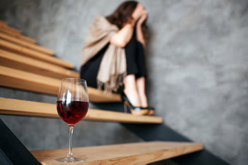 Young woman has problems with alcohol. Sitting on steps and hiding face. Fashionable woman suffer from hangover. Alone in danger. Drunk. Glass of red wine in fron of picture.