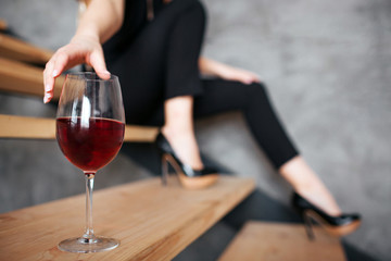 Young woman has problems with alcohol. Cut view of model reaching hand to glass with red wine. Sitting on steps alone. Stylish black female suit.