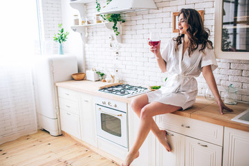 Young woman with dark hair sitting in kitchen and drinking red wine from glass. Enjoying moment. Alone in room. Posing in morning gown.
