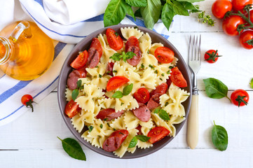 Tasty pasta farfalle with grilled sausages, fresh cherry tomatoes and basil on a plate on a white background. Top view, flat lay.