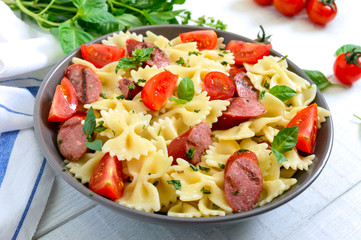 Tasty pasta farfalle with grilled sausages, fresh cherry tomatoes and basil on a plate on a white background.