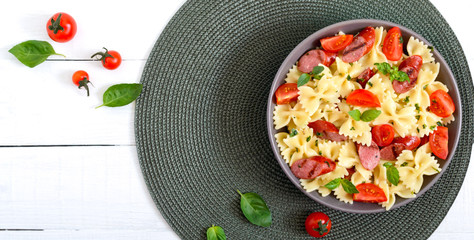 Tasty pasta farfalle with grilled sausages, fresh cherry tomatoes and basil on a plate on a white background. Top view, flat lay. Banner